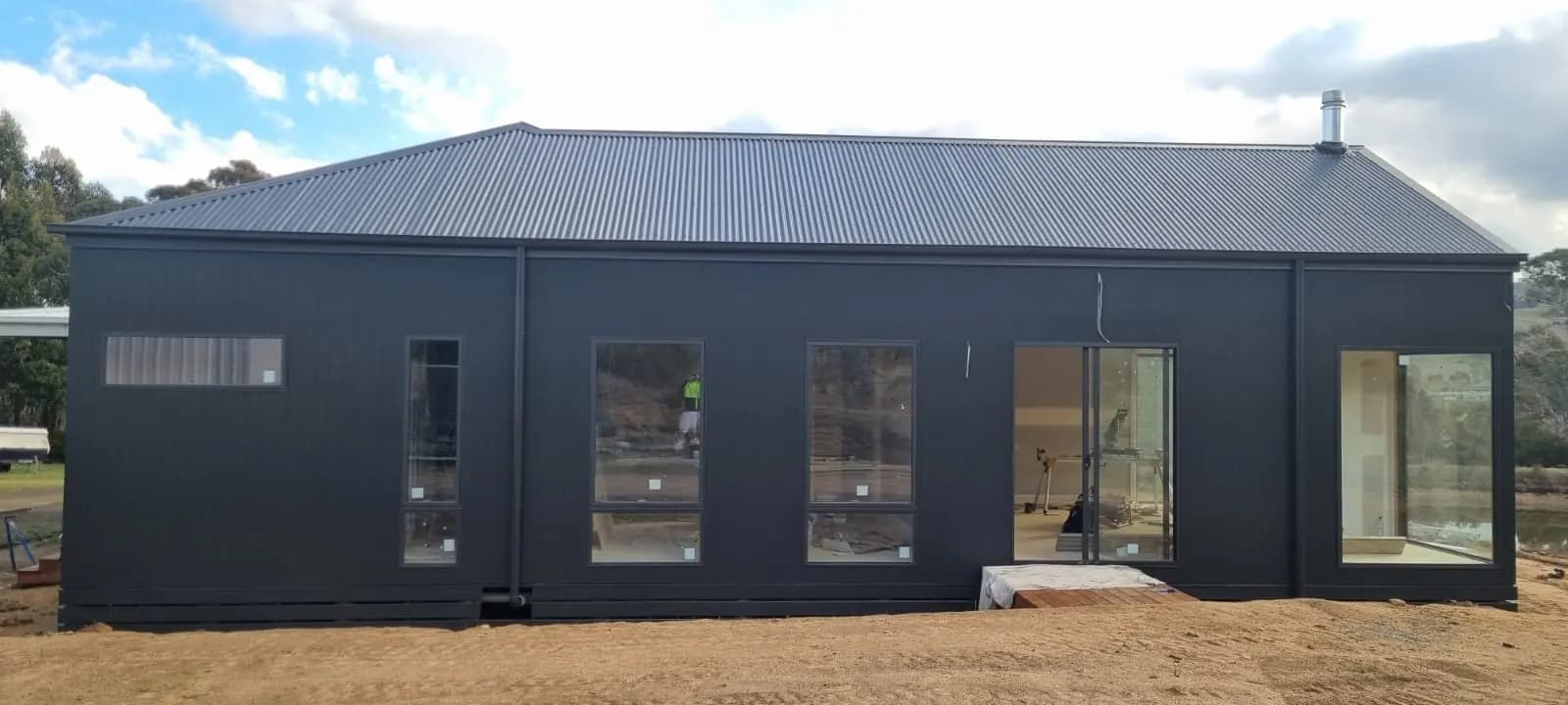 Long elevation of a new extension finished in charcoal cladding under a corrugated steel roof, with a row of black-framed windows and a glazed door.