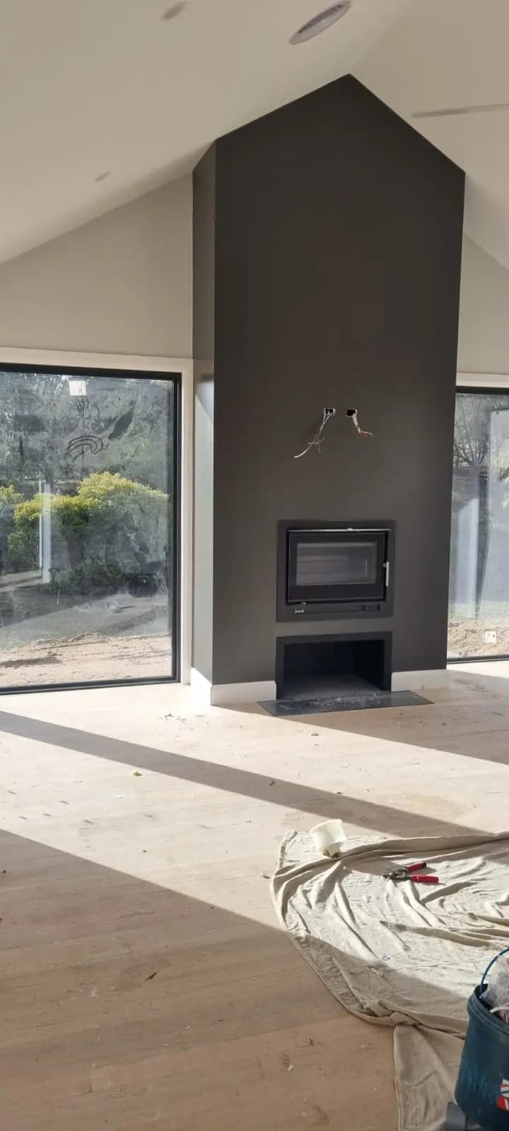 Interior of a new extension with a charcoal-painted chimney breast running to a raked white ceiling, a built-in wood heater, timber floorboards and full-height windows either side.