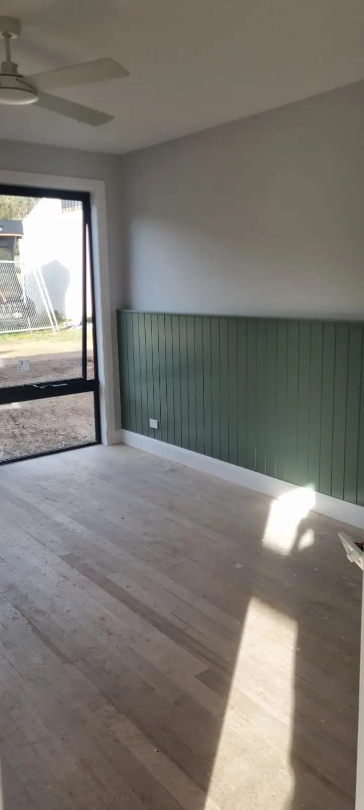 Room in a new extension with green vertical panelling to dado height below pale grey walls, timber floorboards, a ceiling fan and a glazed door to the yard.