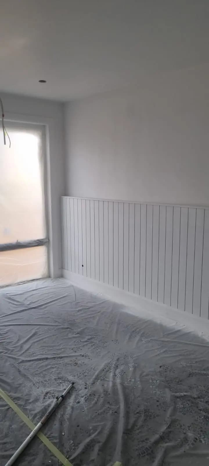 Bedroom in a new extension with white vertical panelling to dado height, white walls above, a masked window and drop sheets across the floor.