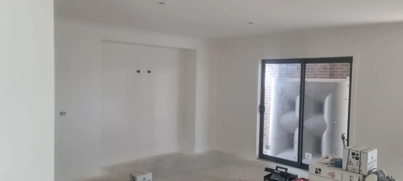 After — Living area of a new house with walls and ceiling painted white, a recessed alcove in the far wall and a black-framed sliding door looking onto a brick courtyard.