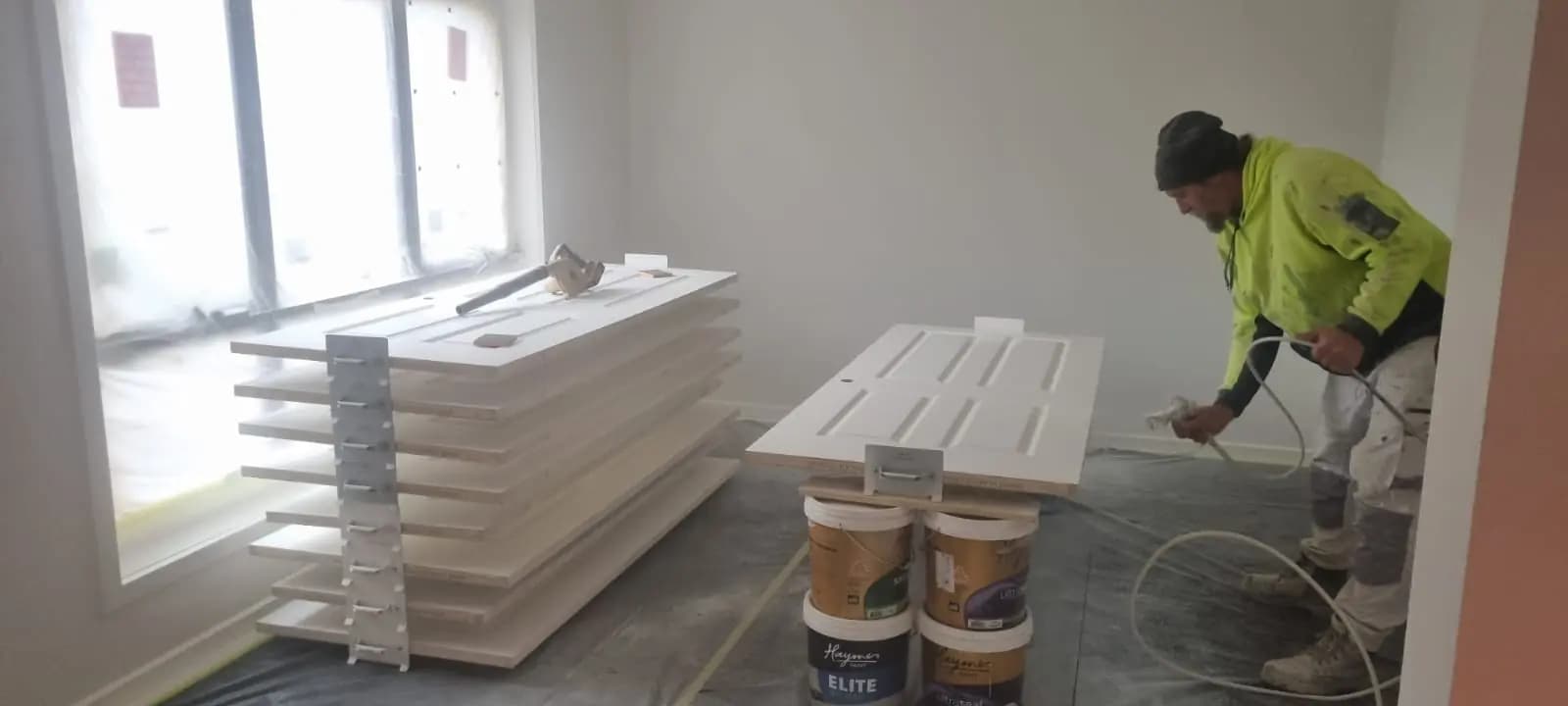 A painter in a hi-vis jumper spray painting a panelled door laid flat, with a rack of freshly sprayed doors and shelves beside him and paint tins on the floor.