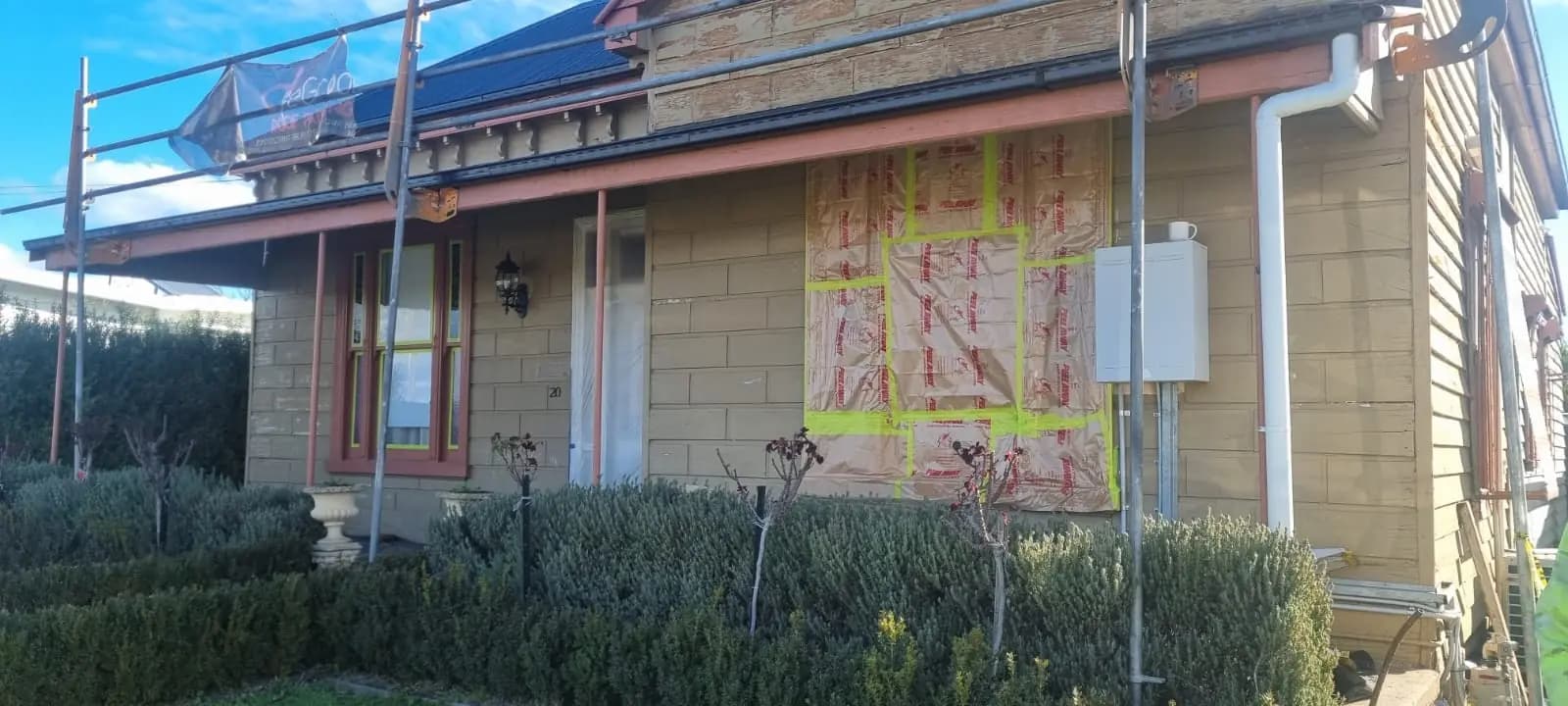 Front of a single-storey weatherboard and rendered-block cottage in faded cream paint, with scaffolding along the wall and a taped sheet of builder's wrap covering a patched section beside the downpipe.