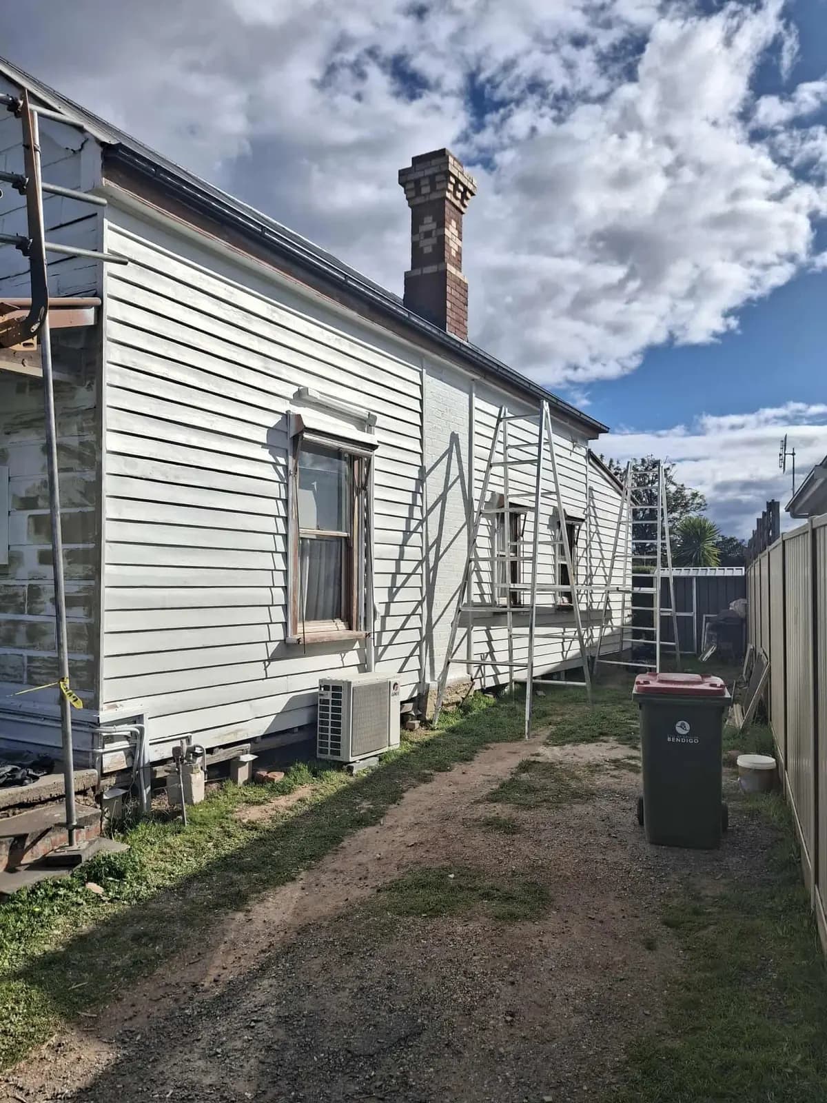 Side elevation of a weatherboard cottage finished in white, with a tall red brick chimney, scaffolding still standing along the wall and a gravel driveway alongside.